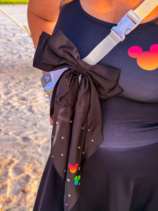 A close-up of a person wearing a dark navy dress with a large black bow accessory tied around the waist, decorated with small silver rhinestones and colorful Mickey Mouse head motifs near the bottom. The background shows sandy beach ground.