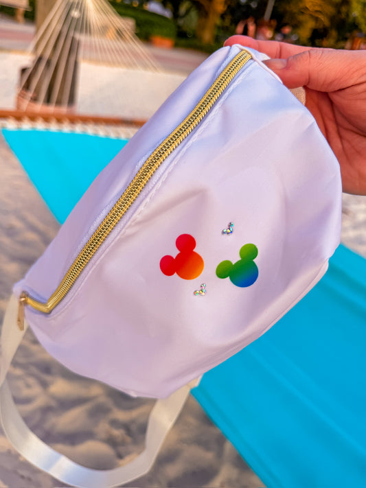 A white nylon fanny pack with a gold zipper, held by a hand over a bright blue beach towel on a sandy beach. The front features two Mickey Mouse-style head silhouettes in a red-to-yellow gradient and a green-to-blue gradient, accented with small rhinestone butterflies.