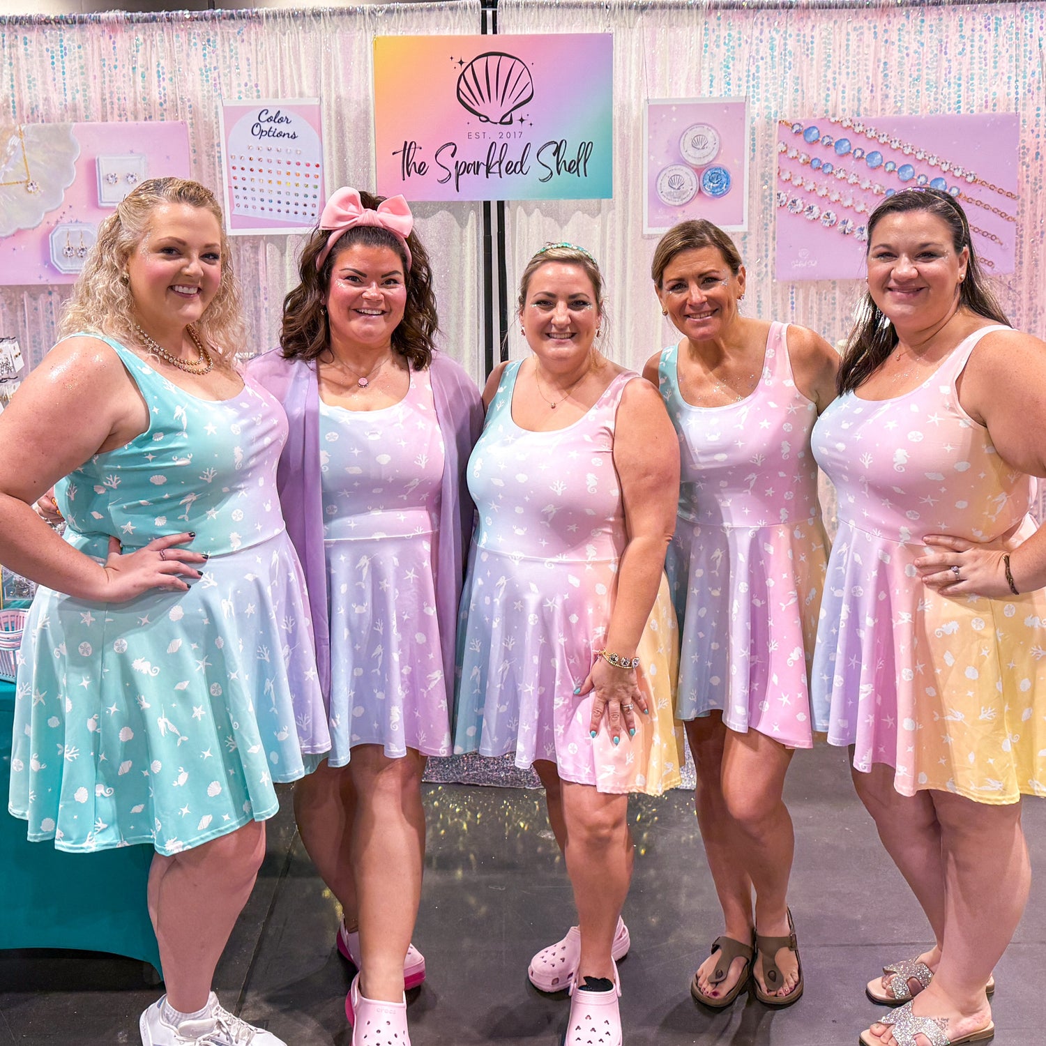 Five women in pastel dresses standing in front of a booth with colorful displays.