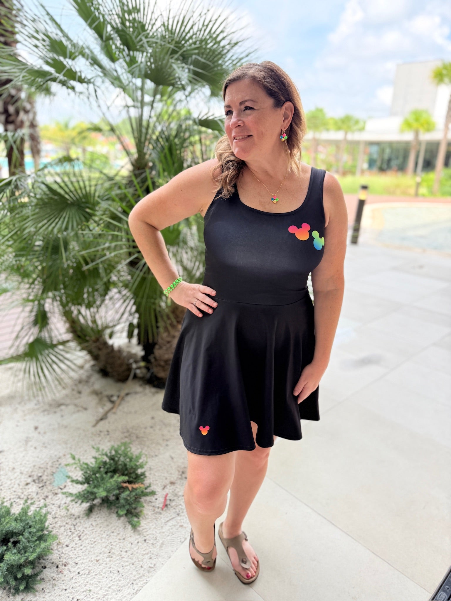 A woman wearing a black sleeveless skater dress with small colorful heart appliqués, standing outdoors on a paved walkway with palm trees and a building in the background.
