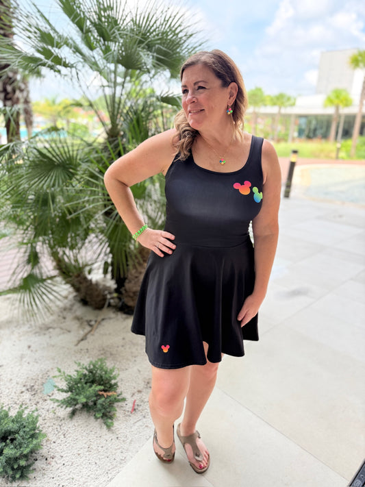 A woman wearing a black sleeveless skater dress with small colorful heart appliqués, standing outdoors on a paved walkway with palm trees and a building in the background.